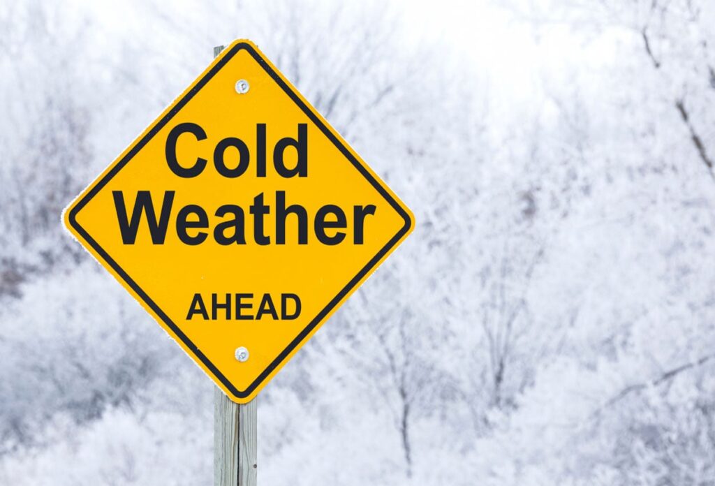 Yellow diamond-shaped warning sign reading 'Cold Weather AHEAD' against a snowy winter background with frost-covered trees.