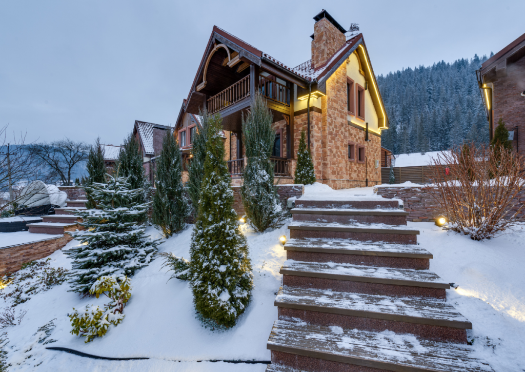 Luxurious brick home with illuminated windows and columned entrance pathway surrounded by snow-covered landscaping and evergreen trees.