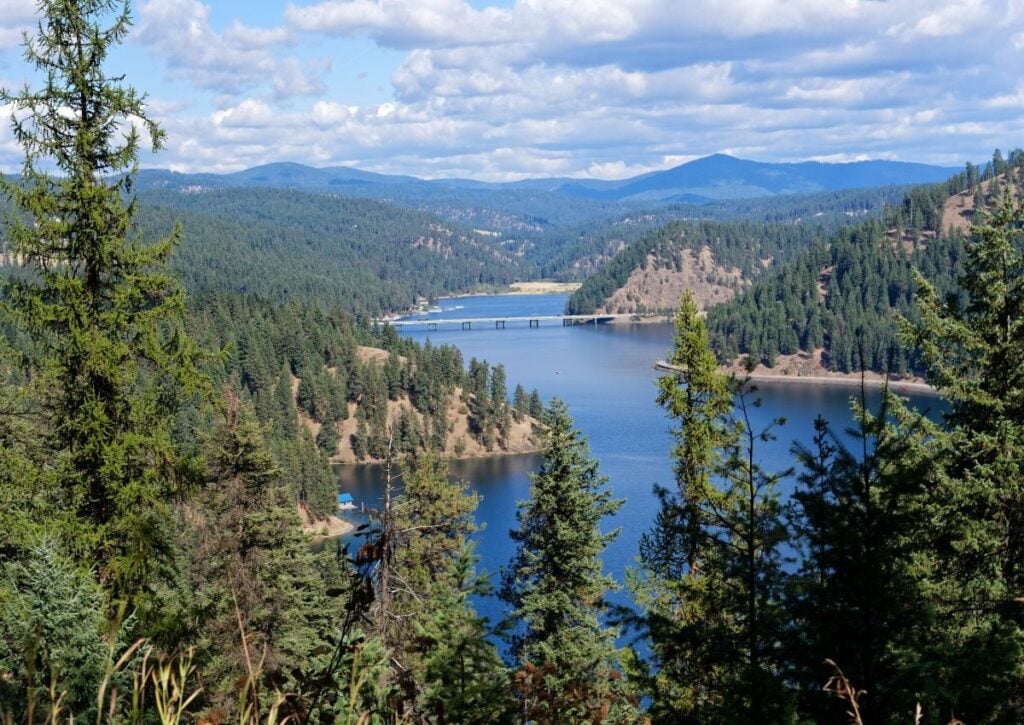 Pristine alpine lake surrounded by dense coniferous forest and mountains under blue sky with white clouds.