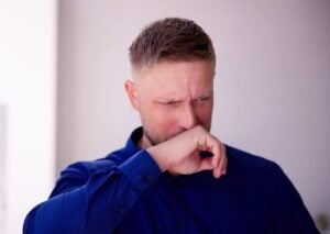 Man in blue shirt coughing with hand to mouth, experiencing discomfort from chemical smell or fumes.