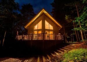 A-frame cabin glowing with warm interior lighting surrounded by tall pine trees in a dark forest setting at dusk.