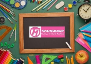Chalkboard displaying Trademark Heating, Cooling & Electrical logo surrounded by school supplies including colored pencils, paintbrush, ruler, compass, and appl