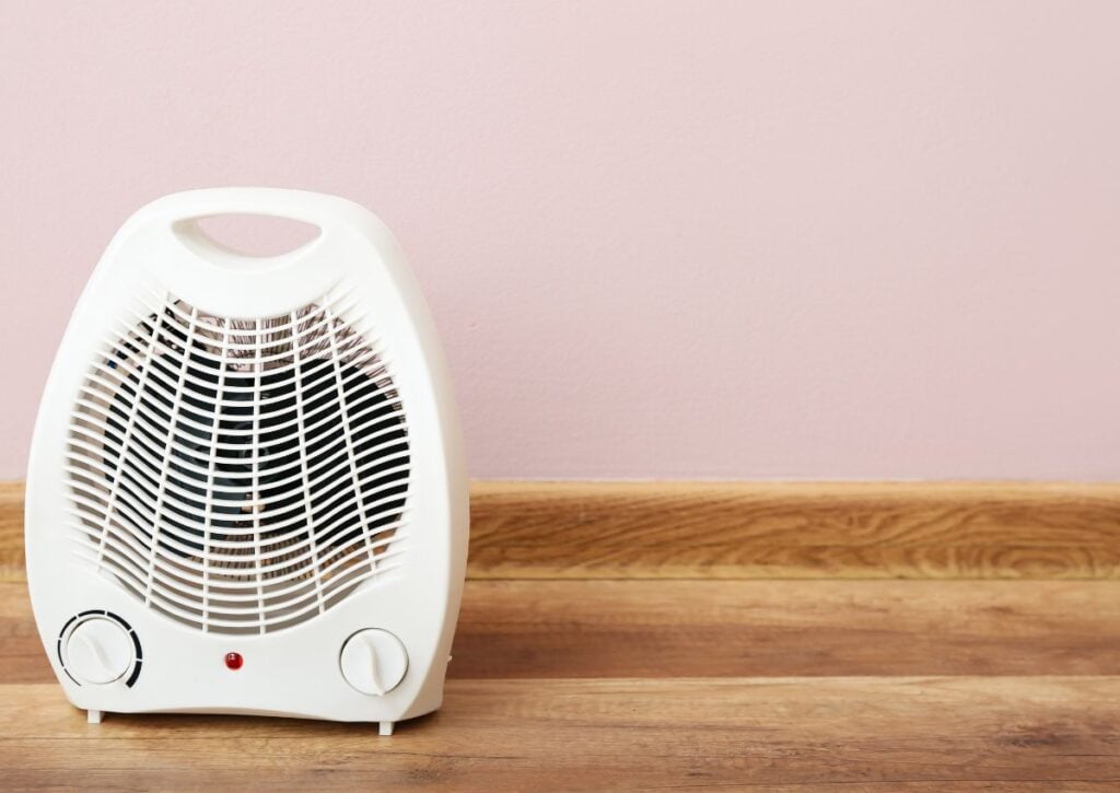 White portable space heater with dual control knobs and indicator light, positioned on wooden surface against pink wall.