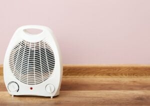 White portable space heater with dual control knobs and indicator light, positioned on wooden surface against pink wall.