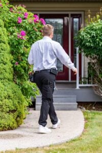Service technician in white shirt and black pants with tool belt jogging toward residential home entrance surrounded by pink flowering bushes.