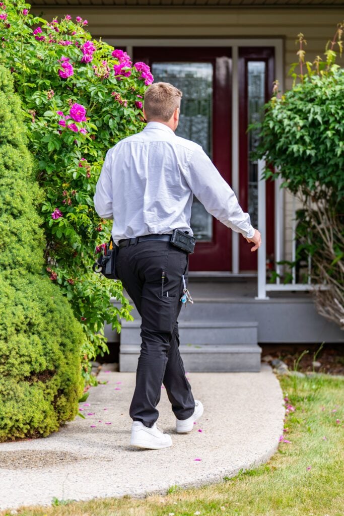 Service technician in white shirt and black pants with tool belt jogging toward residential home entrance surrounded by pink flowering bushes.