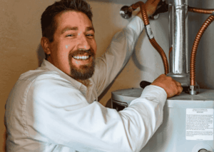 Smiling plumber in white shirt working on water heater installation with copper pipes and valves visible.