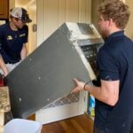 Two technicians installing large metal HVAC ductwork into a residential ceiling during air conditioning system installation.
