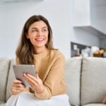 Woman in tan cardigan sits on gray couch while reviewing a document in her hands, appearing thoughtful.