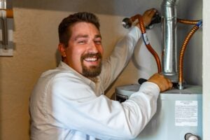Smiling plumber in white shirt installing or servicing a water heater with copper tubing and metal piping visible.